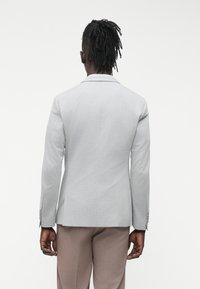 Giacca da completo grigio chiaro con design aderente, spalle strutturate, rever a lancia e due spacchi posteriori. Abbinata a pantaloni beige.