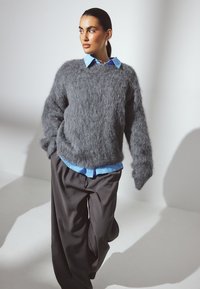 Suéter gris de felpa con mangas largas superpuesto a una camisa de color azul claro con cuello, combinado con pantalones negros holgados. Texturas suaves y materiales agradables.