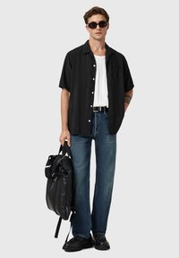 Sort kortærmet skjorte med knapper over en hvid t-shirt, parret med lyseblå jeans og sorte sko, holdende en sort rygsæk.