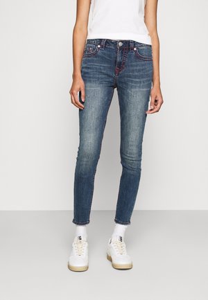 Person, der bærer falmede blå skinny jeans med røde syninger, hvide sokker, hvide sneakers og en ærmeløs hvid top mod en ensfarvet baggrund.