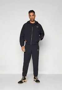 Conjunto atlético preto composto por uma camisola com fecho éclair e calças a condizer; apresenta detalhes com logótipos, punhos elásticos e capuz. Ténis desportivos completam o visual.