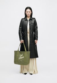 Fekete bőr trench kabát, krémszínű felső és b�ézs pliszírozott nadrág. Zöld bevásárlótáska fehér "Café Kitsuné" logóval.