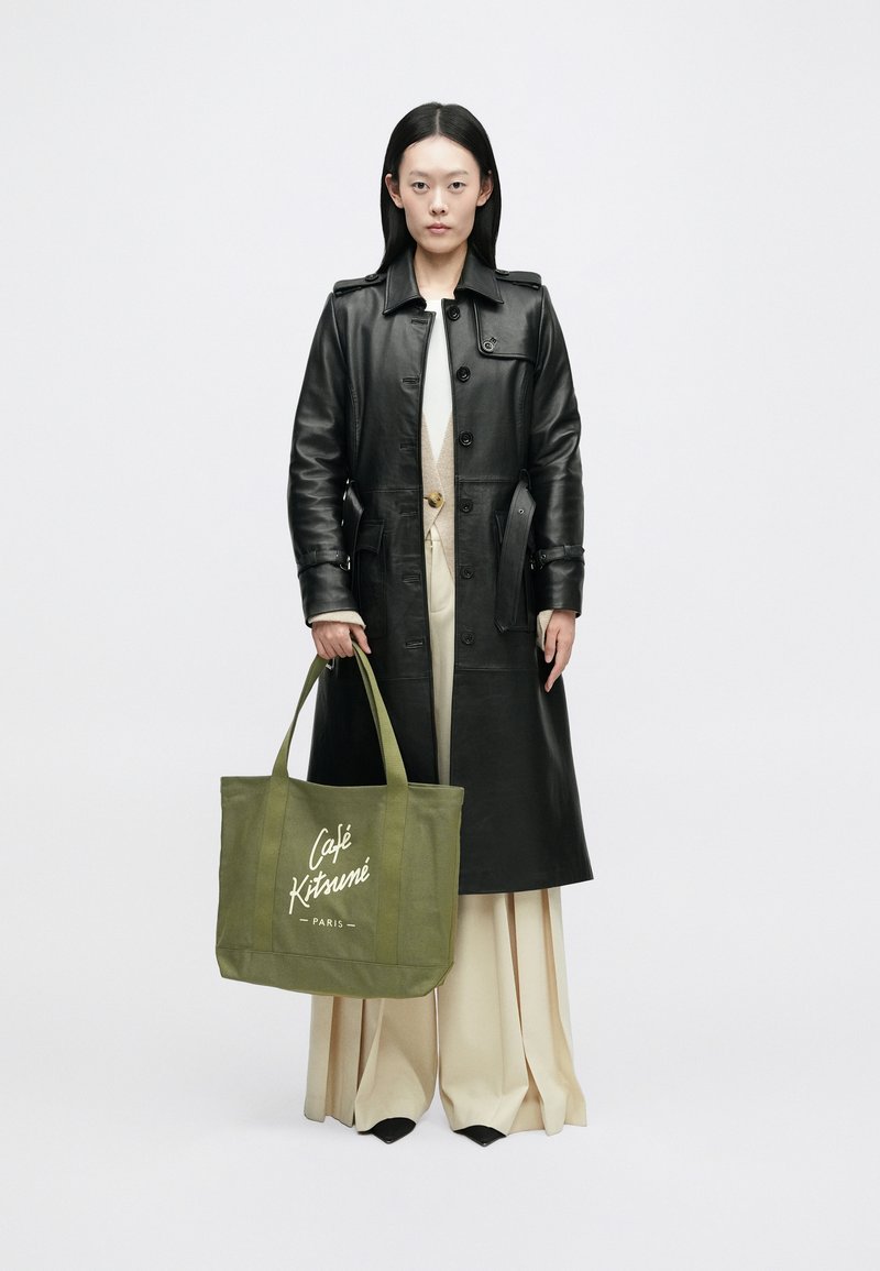 Fekete bőr trench kabát, krémszínű felső és bézs pliszírozott nadrág. Zöld bevásárlótáska fehér "Café Kitsuné" logóval.