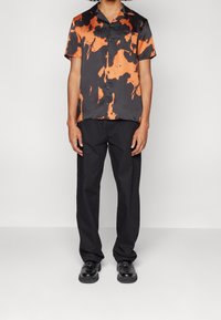 Kurzärmliges Hemd in schwarz-orange abstractem Muster, kombiniert mit schwarzen Hosen und schwarzen Schuhen, was ein kontrastierendes Ensemble schafft.