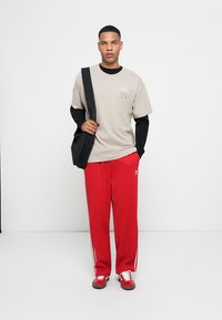 Lys grå t-shirt med logo, sort langærmet skjorte under, røde træningsbukser med hvide striber og røde sneakers. Sort skuldertaske.