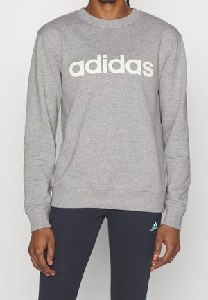 Osoba oblečená v světle šedé mikině Adidas a tmavých legínách s zeleným logem Adidas na stehně, stojící před jednobarevným pozadím.