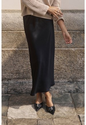 Schwarzer Maxirock aus Satin, kombiniert mit einem strukturierten, beige gestrickten Pullover. Dazu schwarze, spitz zulaufende Pumps mit dekorativen Schnallen.