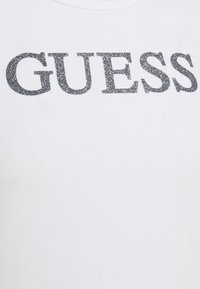 Camiseta blanca con la palabra "GUESS" en letras negritas y brillantes de color plata. La tela parece suave y presenta un diseño estándar de cuello redondo.