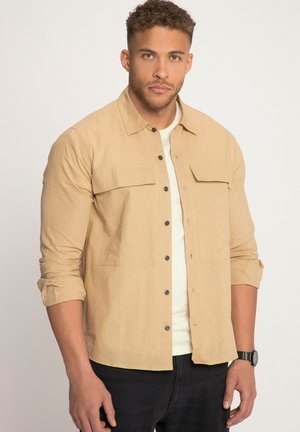 Helles, beige Hemd mit zwei Brusttaschen, Knopfverschluss, aufgerollten Ärmeln und glatter Oberfläche. Getragen über einem weißen T-Shirt und schwarzen Jeans.