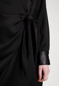 Robe noire avec un détail de nouage à la taille, manches longues avec un poignet en cuir, et un tissu doux et texturé.