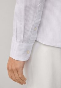 Linke Hand einer Person, die ein weißes und hellgrau gestreiftes Langarmhemd mit geknöpften Manschetten trägt, entspannt an ihrer Seite.