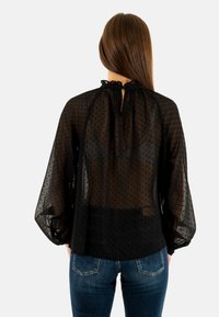 Blusa negra transparente con lunares, mangas largas abullonadas, cuello con lazo y un ajuste ligeramente holgado. Llevada sobre jeans oscuros.