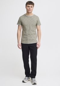 Tricou scurt de culoare verde măsliniu, cu mânecă scurtă și tiv alb, combinat cu pantaloni negri și adidași gri, stând în fața unui fundal simplu.