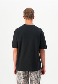 Bărbat cu păr scurt și ondulat, purtând un tricou negru și pantaloni bej cu un model, stând cu spatele într-un fundal alb.