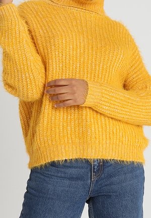 Personne portant un pull tricoté épais jaune vif à manches longues et un jean bleu sur un fond clair uni.