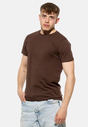 Braunes Baumwoll-T-Shirt mit Rundhalsausschnitt und kurzen Ärmeln, das ein figurnahes Design aufweist. Kombiniert mit hellblauen Jeansshorts.