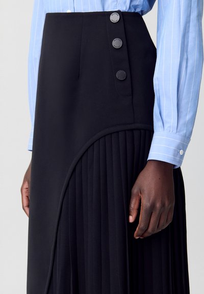 Gonna nera plissettata con una cintura liscia che presenta tre bottoni neri rotondi. La gonna ha un dettaglio di cucitura curva. Camicia a righe blu chiaro visibile.