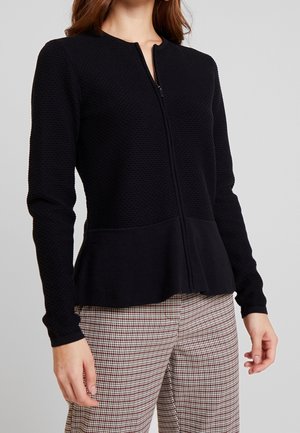 Kobieta w teksturowanej czarnej kurtce zapinanej na zamek z peplum, nosząca brązowo-białe spodnie w kratę, stojąca na tle jednolitego szarego tła.