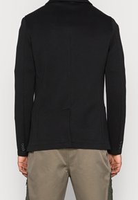 Svart figursydd kavaj i slätt tyg, med slagkrage, strukturerade axlar och två knappar på varje ärm.