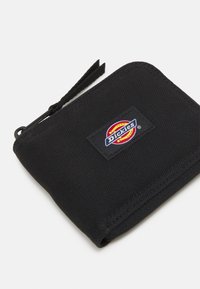 Cartera negra con cremallera hecha de tejido texturizado, con un destacado parche de logo rectangular que presenta una colorida marca.