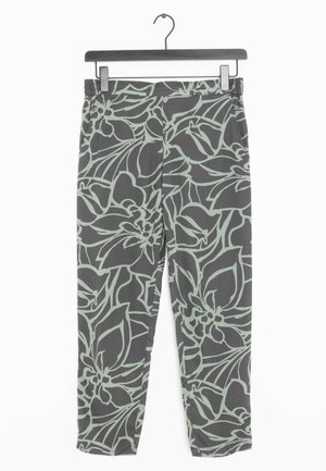 Ciemnoszare spodnie z jasnym zielonym, abstrakcyjnym wzorem kwiatowym zawieszone na czarnym, drewnianym wieszaku na białym tle.