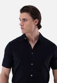 Camicia corta blu scuro con colletto, dotata di una pattina con bottoni grigi e un tessuto liscio e texture.