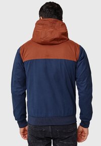 Homme portant une veste à capuche bleu marine et marron rouille, vu de dos, avec un jean noir sur un fond gris uni.