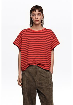 Joven con cabello corto que lleva una camiseta de rayas rojas y negras de gran tamaño y pantalones marrones holgados frente a un fondo blanco liso.