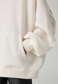 Felpa crema con una vestibilità rilassata, polsini a coste e taschino anteriore. Caratterizzata da borchie sulla spalla sinistra per un tocco di dettaglio. Texture morbida.