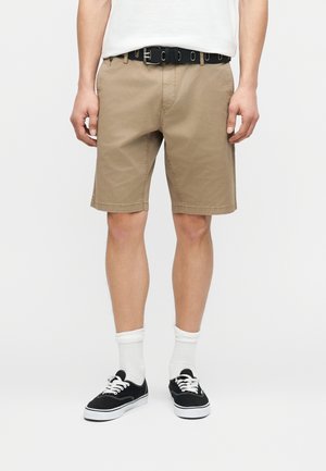 Mann trägt beige kniehohe Shorts, ein weißes Hemd, das in die Shorts gesteckt ist, einen schwarzen Gürtel, weiße Kniestrümpfe und schwarze Schnürschuhe, steht vor einem weißen Hintergrund.
