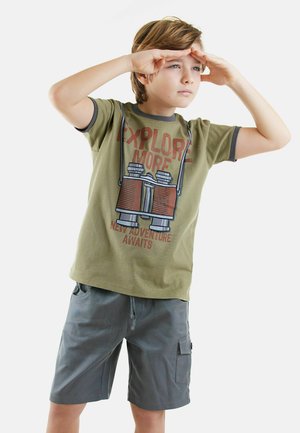 Grøn T-shirt med "Explore More" tryk og kikkertgrafik; grå shorts med lommer; dreng der beskytter sine øjne, lys baggrund.