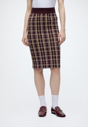 Donna che indossa una gonna a quadri bordeaux e beige al ginocchio, calzini bianchi alla caviglia e mocassini bordeaux su sfondo uniforme.