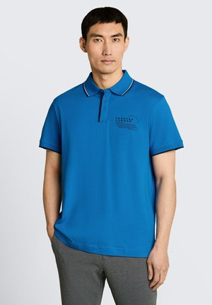 Mężczyzna w niebieskiej koszulce polo z napisem „Forever Summer” i szarych spodniach, stojący z jedną ręką w kieszeni na jednolitym tle.