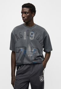 Σκούρο γκρι φαρδιά μπλούζα με μεγάλο γραφικό εκτύπωμα που περιλαμβάνει αριθμούς και ένα στιλιζαρισμένο λογότυπο. Κοντά μανίκια, λαιμόκοψη με στρογγυλή γραμμή, απαλό βαμβακερό ύφασμα.