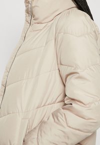Světlé béžové péřové sako s předním zipem, prošívaným designem a vysokým límcem. Tkanina má hladkou, lesklou texturu.