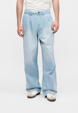 Homme portant un jean bleu clair à jambes larges avec plis avant et des baskets blanches, debout sur un fond blanc uni.