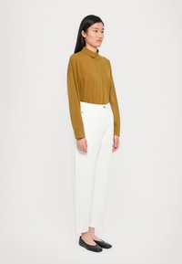 Camicetta a maniche lunghe color senape con colletto abbottonato, abbinata a pantaloni bianchi a vita alta e scarpe nere basse. Design semplice e su misura.