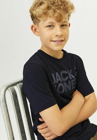 T-shirt preta de manga curta com um grande logo em cinza escuro. Rapaz sentado numa cadeira de metal, com os braços cruzados e cabelo encaracolado.