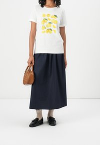 Weißes Kurzarm-T-Shirt mit gelbem Zitronenmotiv, kombiniert mit einem langen marineblauen Rock. Hält eine braune, strukturierte Handtasche. Schwarze Schuhe und beige Socken.