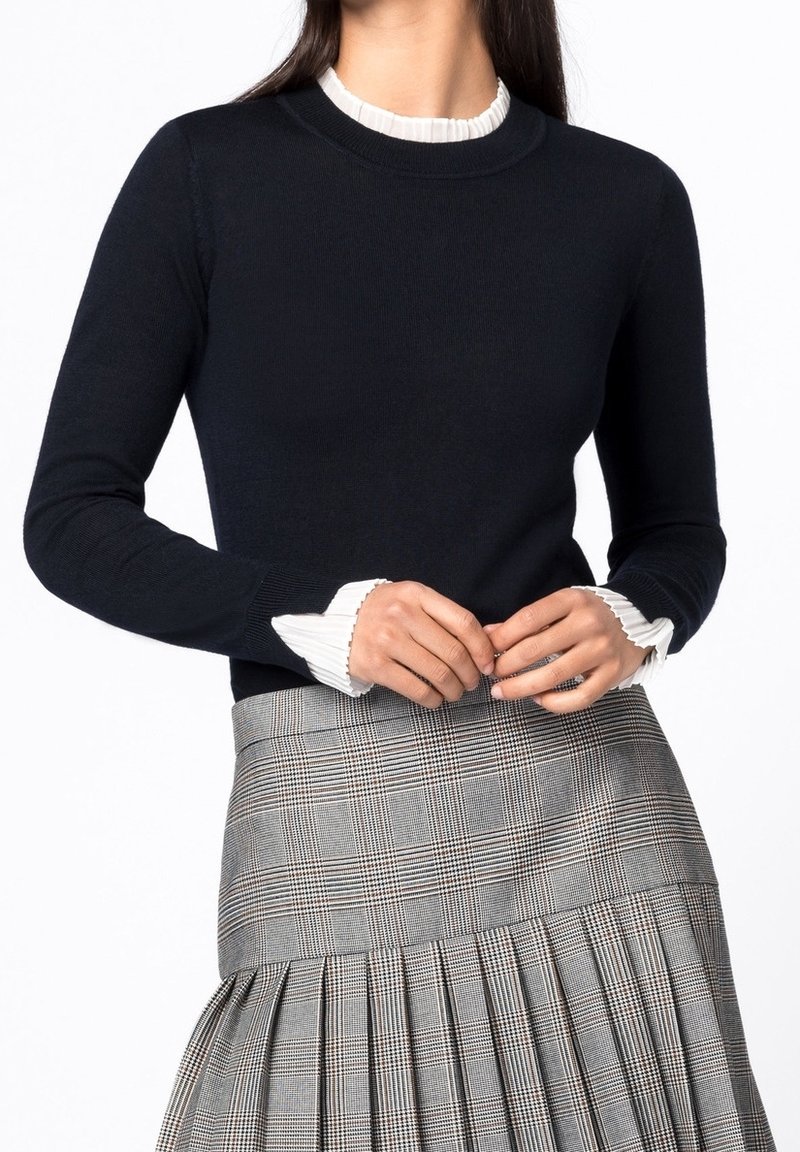 Femme en pull noir avec poignets et col à volants blancs, portant une jupe plissée grise à carreaux, les mains jointes à la taille.