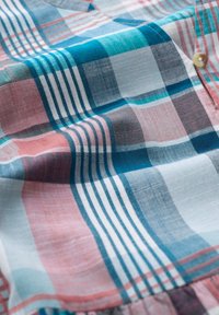 Zbliżenie na pomarszczony materiał w kratę w odcieniach teal, różowego, szarego i białego z jasnobeżowym guzikiem blisko krawędzi.