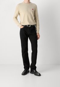 Beige Langarmpullover mit kleinem rotem Logo, kombiniert mit schwarzen Jeans und schwarzen Dressschuhen, vor einem schlichten Hintergrund stehend.