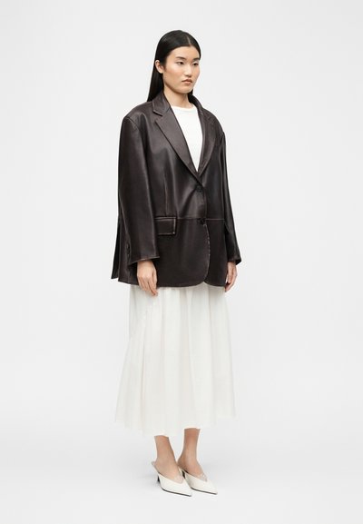 Γυναίκα που φοράει υπερμεγέθη σκούρο καφέ δερμάτινο σακάκι πάνω από λευκό φόρεμα με λευκές μυτερές γόβες slingback σε λευκό φόντο.