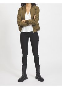 Femme debout avec les bras croisés, portant une veste bomber olive, un chemisier blanc, un pantalon noir, des bottes noires et des colliers superposés sur fond blanc.