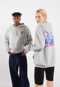 Grå hættetrøjer med frontlomme og farverig grafik på ryggen. Model iført mørke jeans og en anden i sorte shorts. Neutral baggrund.