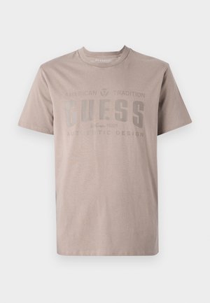 Camiseta de color beige claro, fabricada en algodón, con cuello redondo, mangas cortas y un diseño impreso con el nombre de la marca "GUESS" en gris oscuro.