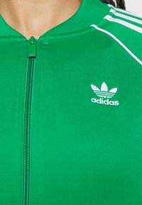 Grön zip-up jacka av slät tyg med vita tre-stripes detaljer och en vit broderad Adidas-logotyp på vänster bröst.