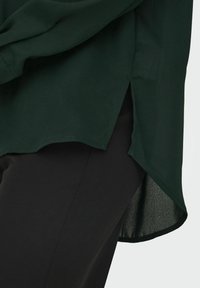 Blusa verde scuro con orlo curvo, fessure laterali e una texture liscia, indossata sopra pantaloni neri dal design semplice.
