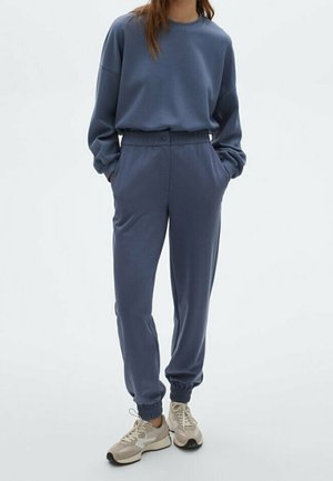 Femme portant un sweat bleu ample et un pantalon de jogging assorti avec des poches, associé à des baskets beige, debout les mains dans les poches.