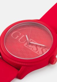 Crvena silikonska sat sa teksturiranim brojčanikom na kojem se ponavlja logo "GUESS", bijelim kazaljkama za sate i minute, te nazubljenim okvirom.